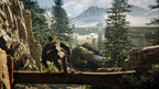 Person sitting on a log in a mountainous landscape with a building and trees in the background